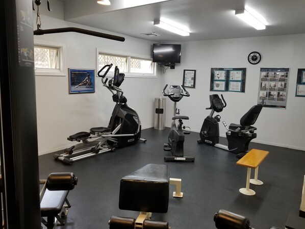 A gym with various exercise equipment, including treadmills, stationary bikes, and weight machines. There are posters on the walls, a clock, and a television mounted on the wall. - Tamarack Beach Resort Hotel (Carlsbad)