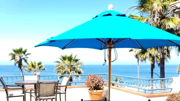 An outdoor patio with round tables and chairs, a blue umbrella, and a view of the ocean. - Tamarack Beach Resort Hotel (Carlsbad)
