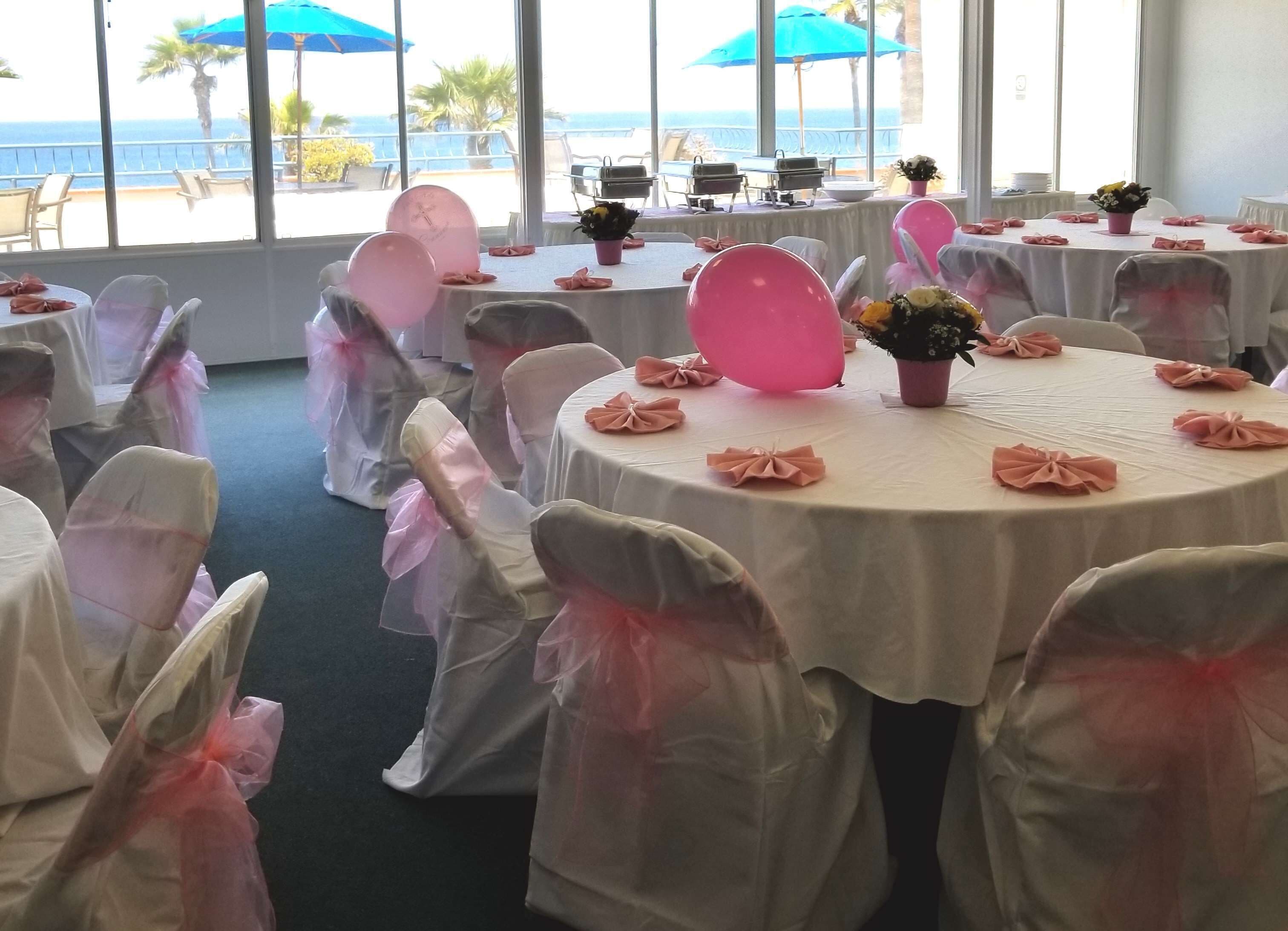A room with round tables set for a formal event, chairs with pink sashes, and a view of the ocean through large windows.