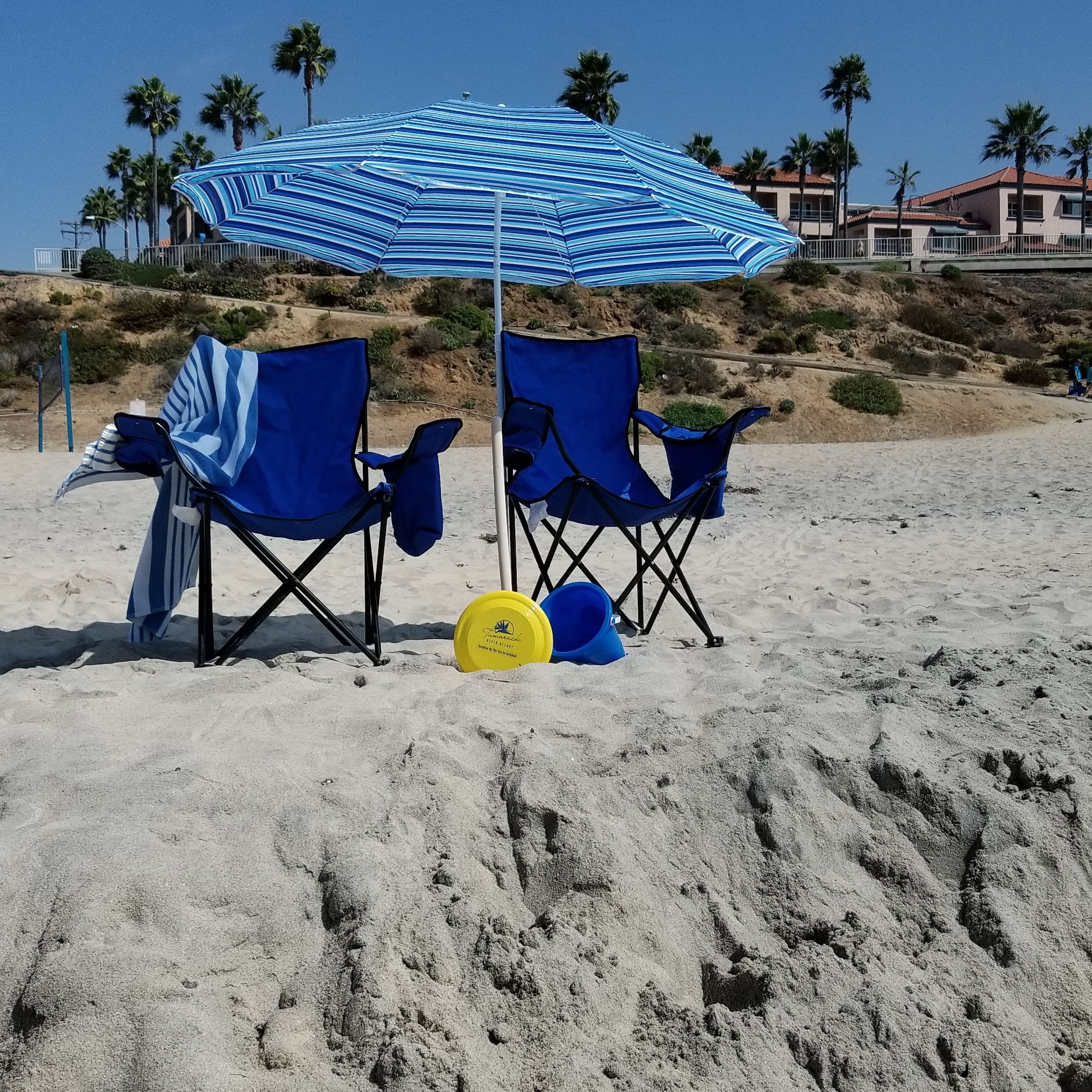 Deux chaises de plage bleues sont placées sous un parasol rayé, sur une plage de sable bordée de palmiers, avec un bâtiment visible au loin.