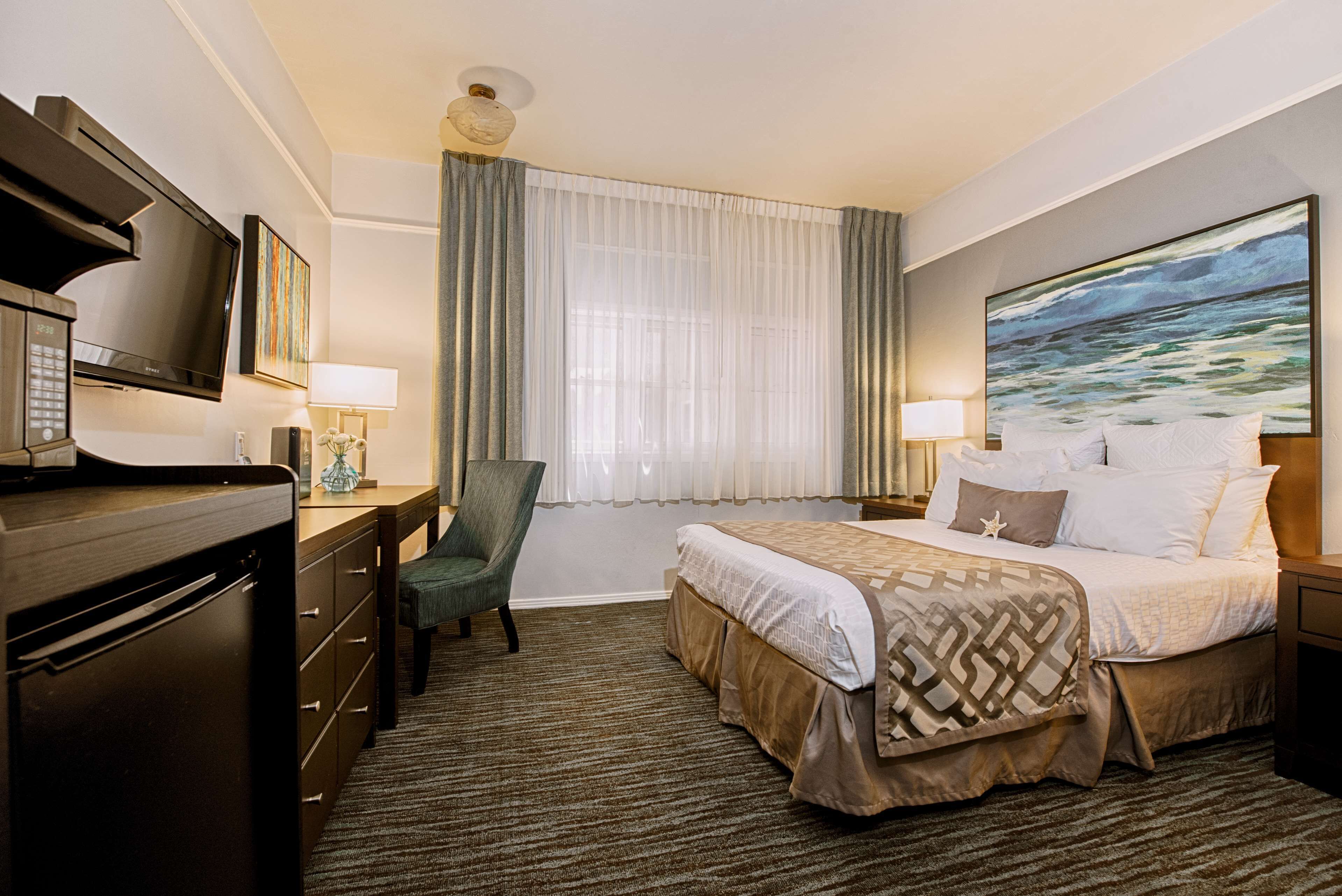 A hotel room with a bed, a desk with a television, a chair, a window with curtains, and a painting on the wall.