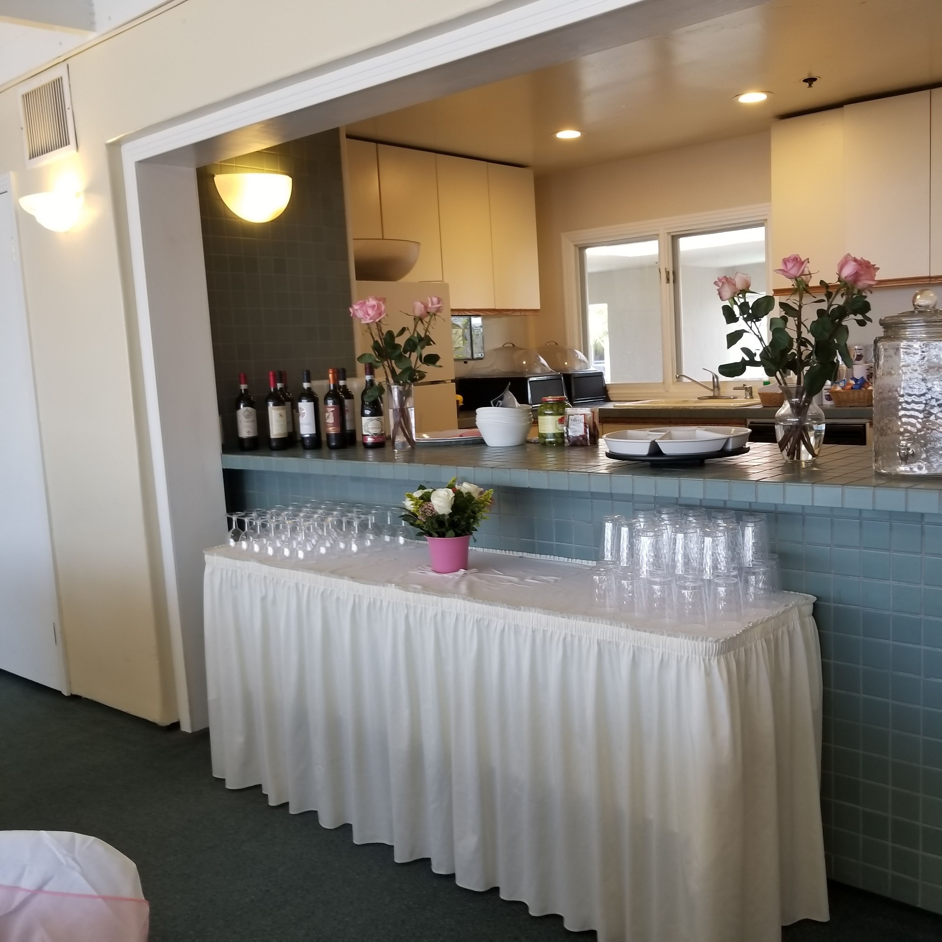 A kitchen area with a counter displaying wine bottles, a vase with flowers, and a table covered with a white cloth.