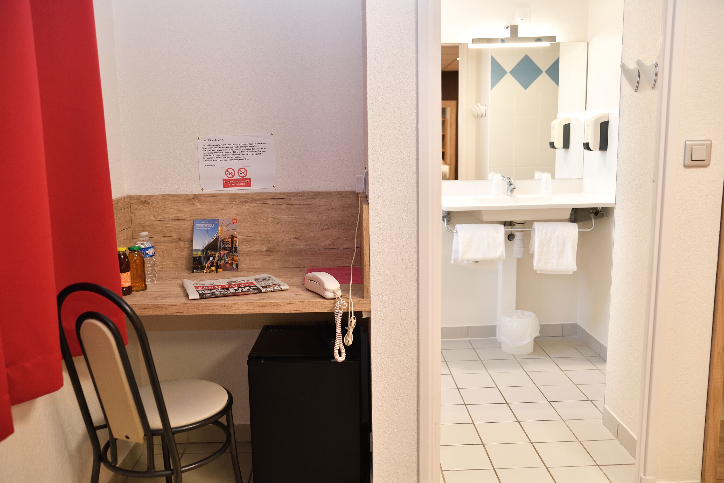 Une salle de bain d’hôtel avec un lavabo, un miroir et une petite poubelle. On y trouve également une chaise et un bureau avec un téléphone et un magazine.