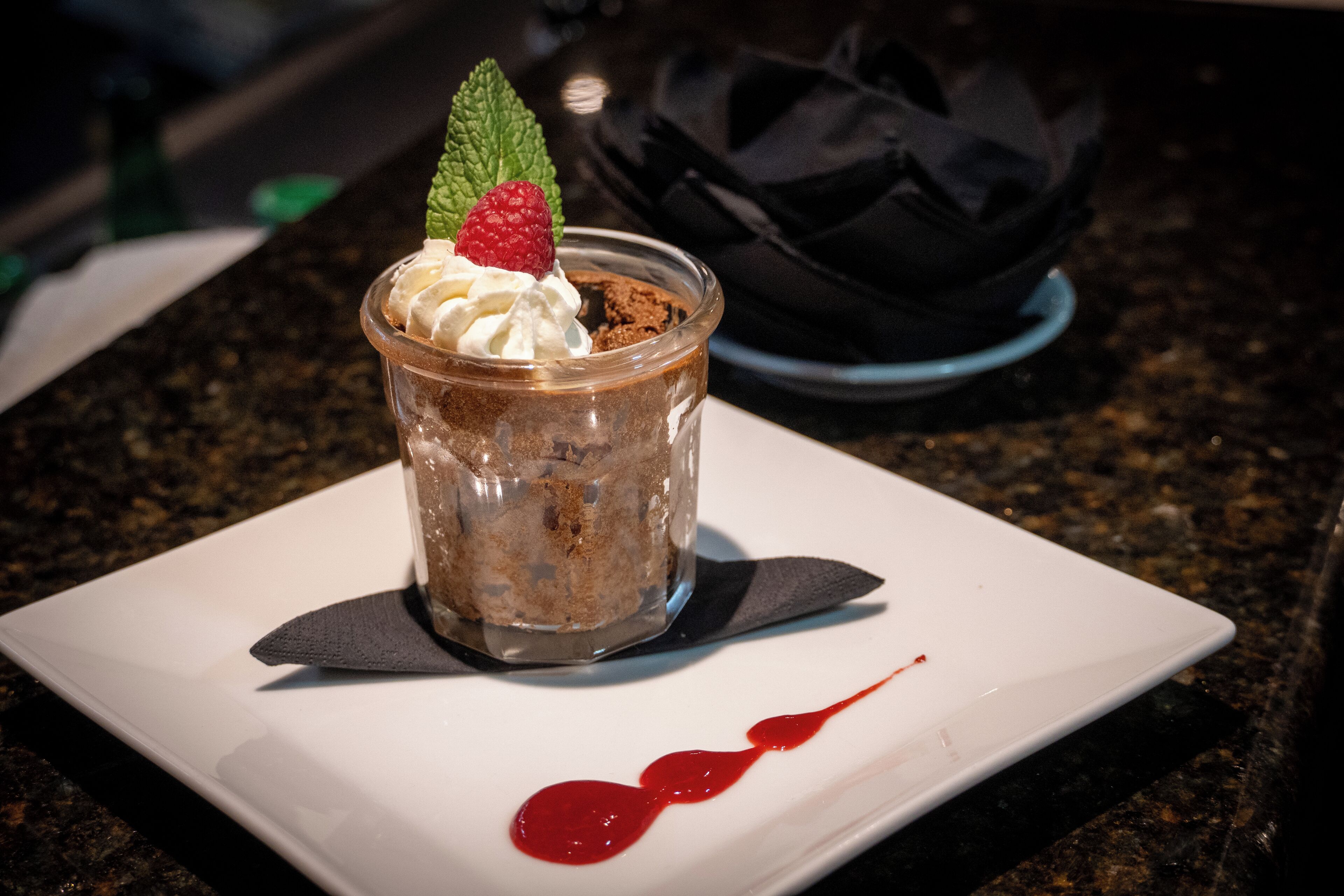 Un postre en un vaso transparente sobre un plato blanco, con un goteo de salsa roja, sobre una encimera de granito oscuro.