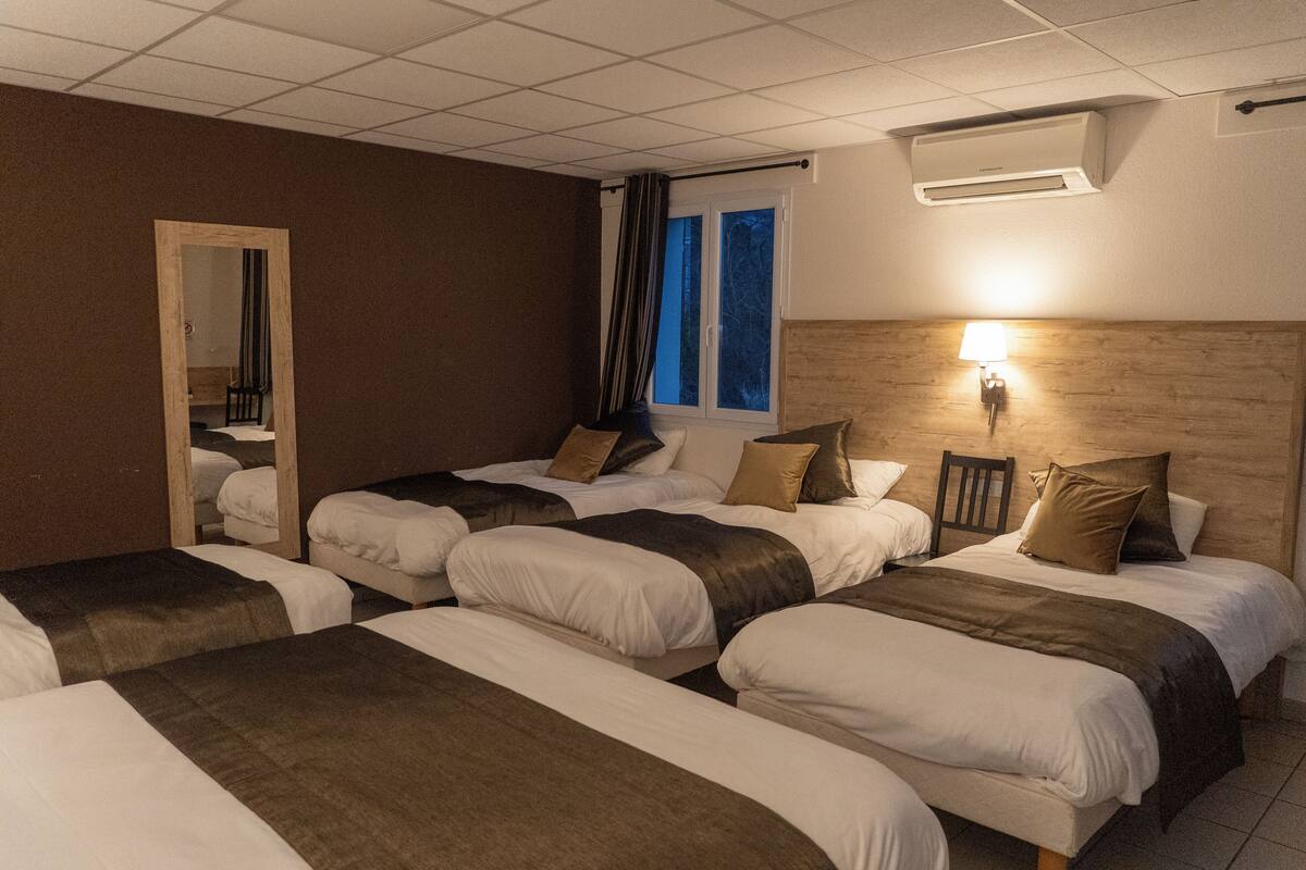 Ein Hotelzimmer mit vier Einzelbetten, die in einer Reihe angeordnet sind, einem hölzernen Kopfteil, einem Nachttisch mit Lampe und einem Fenster mit Vorhängen.