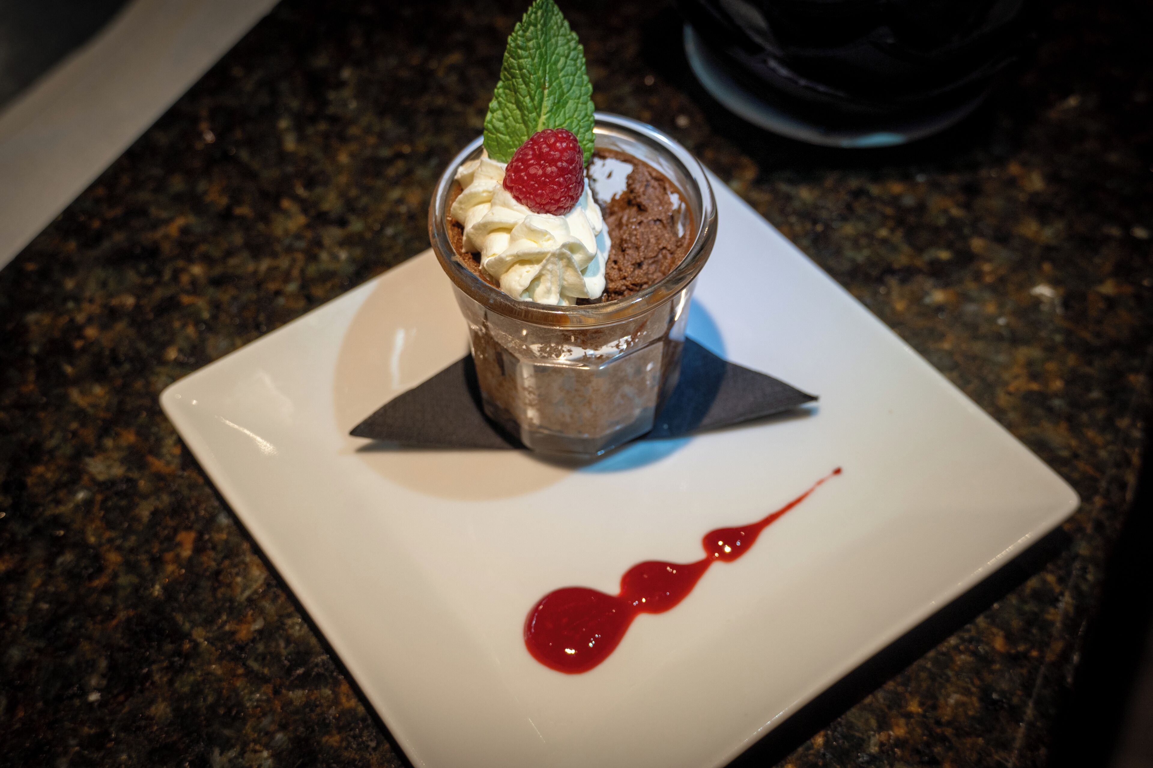 A dessert in a glass cup on a white plate with a red sauce drip.