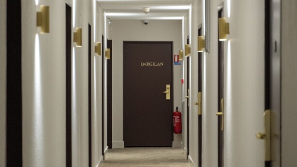 Un couloir d’hôtel avec des portes des deux côtés, un extincteur sur la droite et, au bout, une porte portant l’inscription « DARGILAN ».