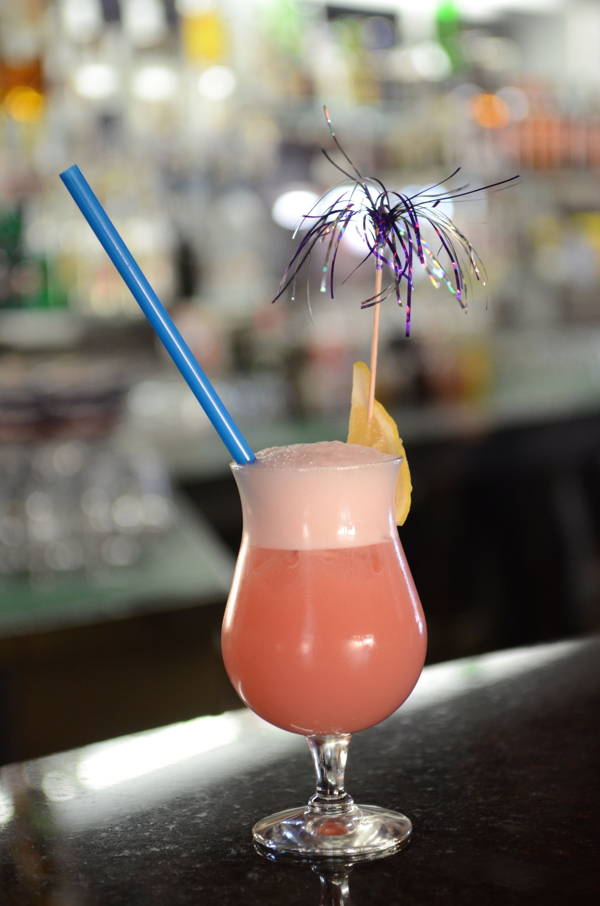Un cocktail avec une paille bleue et un petit parasol décoratif, posé sur le comptoir d’un bar.