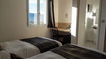 Une chambre d’hôtel avec deux lits, une fenêtre avec des rideaux, un bureau avec une chaise et une salle de bain visible à travers une porte ouverte.