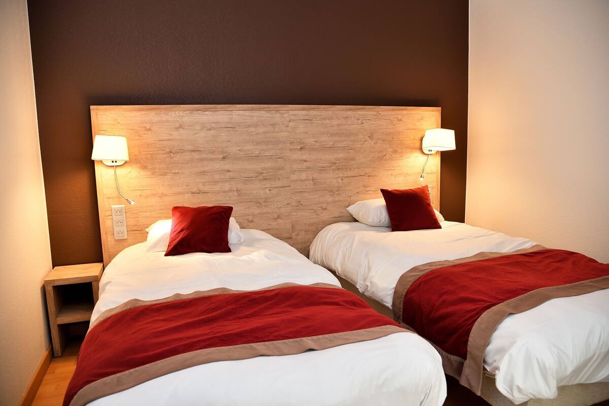 Ein Hotelzimmer mit zwei Betten, jeweils mit Holz-Kopfteil, weißer Bettwäsche und roten Akzenten. Es gibt Nachttischlampen und einen Nachttisch mit einer Lampe.