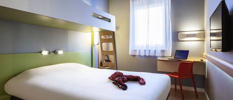 Una habitación de hotel con cama, escritorio, laptop, silla, televisor y ventana con cortinas.