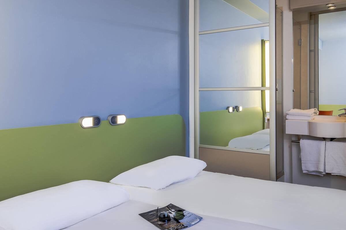 Habitación de hotel con cama, mesita de noche, espejo y baño con lavabo y perchero para toallas.