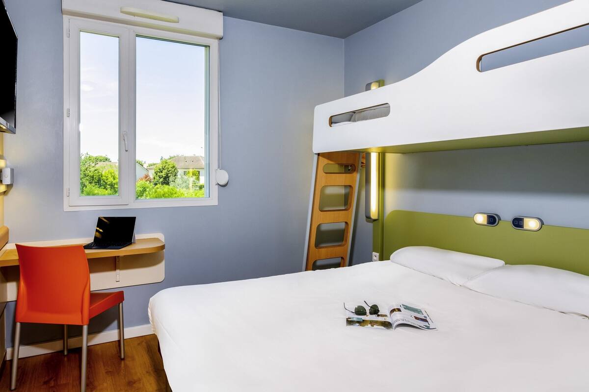 A bunk bed with a ladder, a desk with a laptop, and a window with a view of greenery.