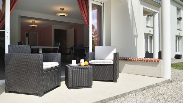 Un área de estar al aire libre con mobiliario de mimbre y una mesa, un porche cubierto y un edificio con columnas blancas y base de ladrillo rojo.