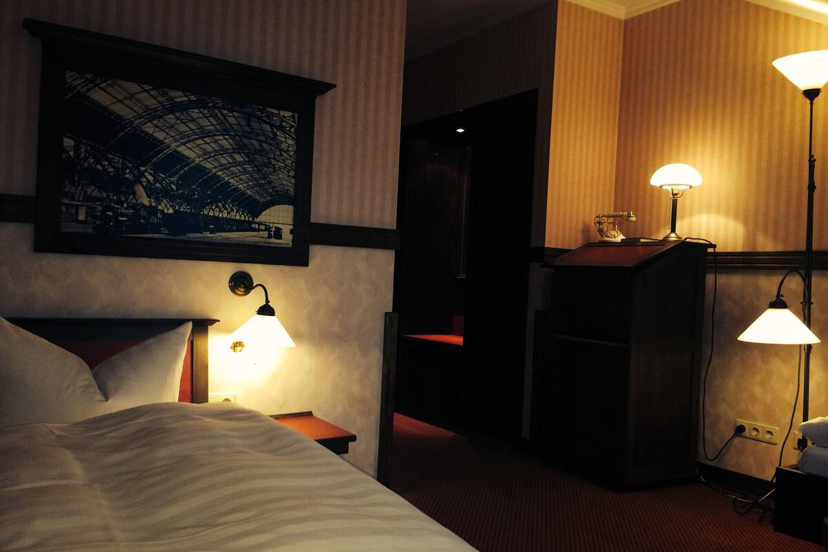 A hotel room with a bed, two bedside lamps, a framed picture on the wall, and a desk with a lamp.