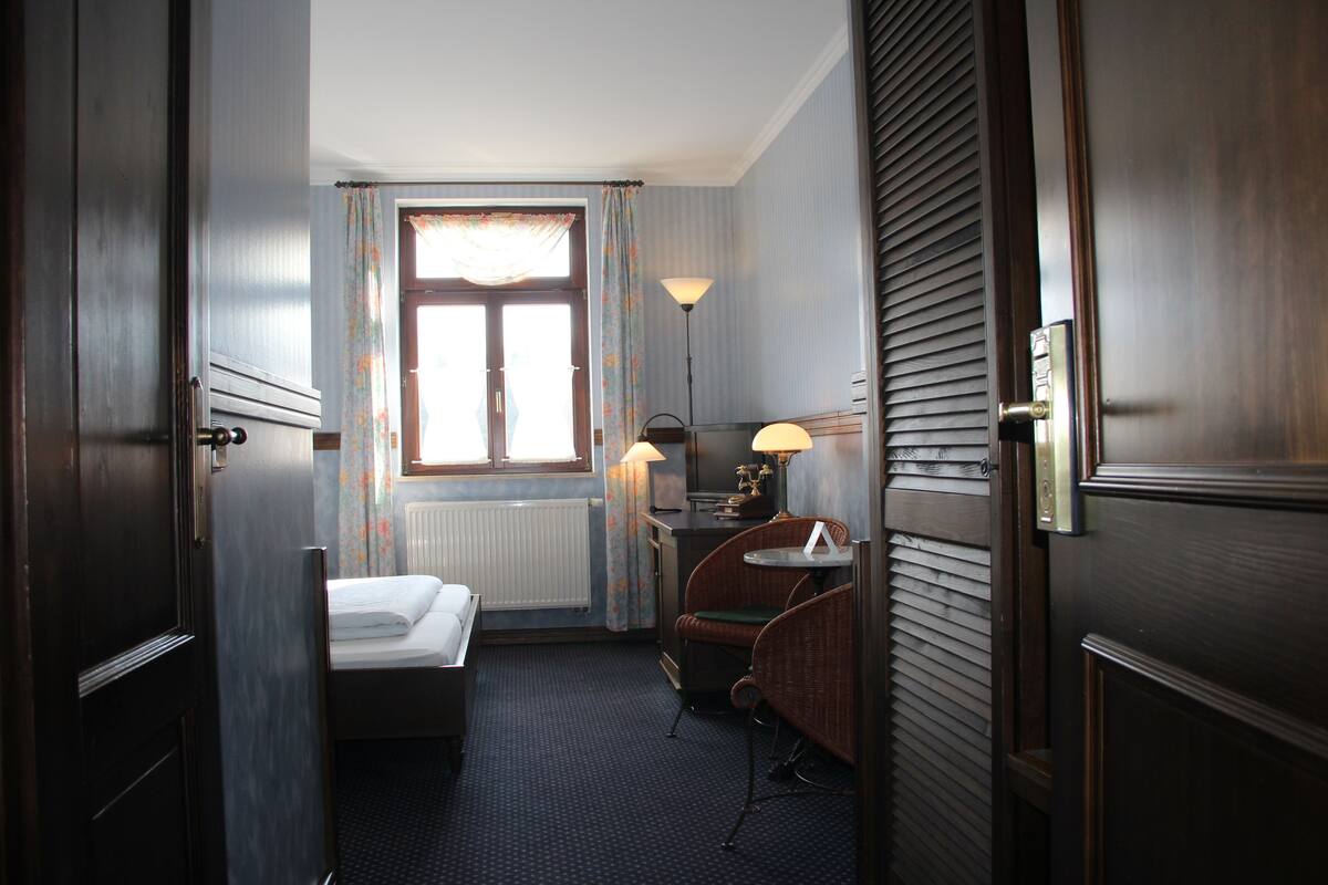 A hotel room with a bed, a small table with a lamp, a chair, and a window with curtains.