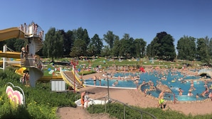 Een groot buitenzwembad omringd door een park met bomen en een waterglijbaan.