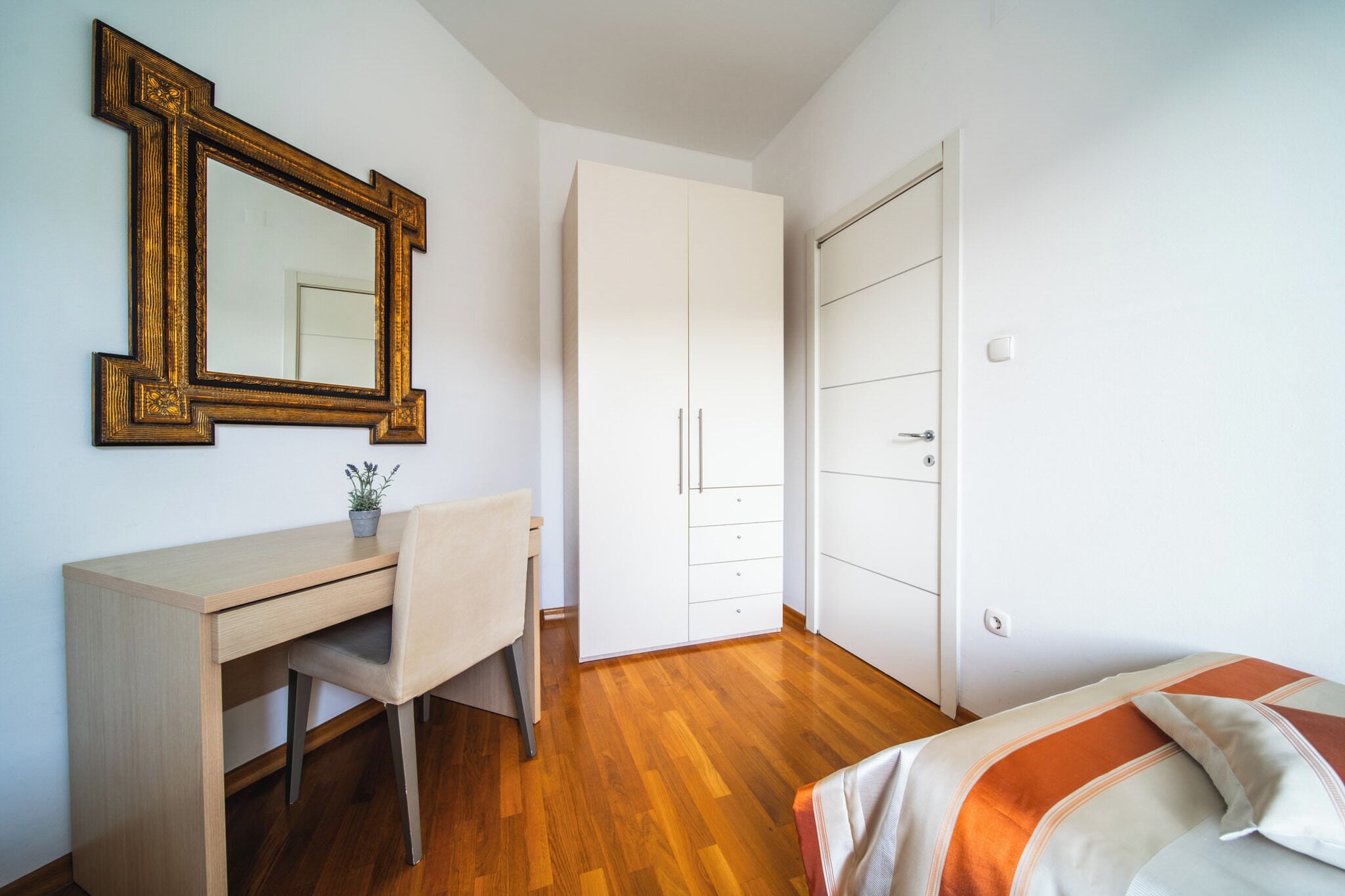 Un dormitorio con escritorio de madera, una silla, cama con ropa de cama a rayas, armario y un espejo en la pared.