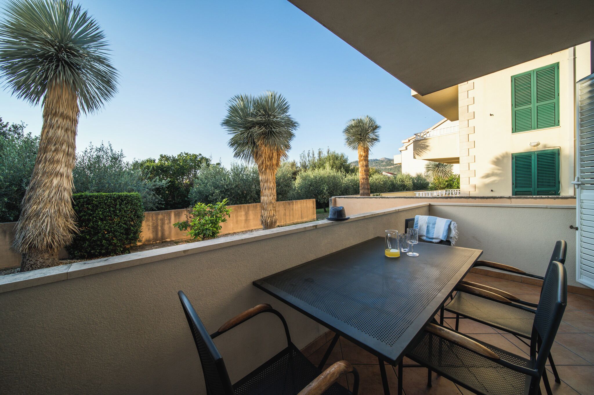 A balcony with a table and chairs, surrounded by palm trees and a view of a building with green shutters.