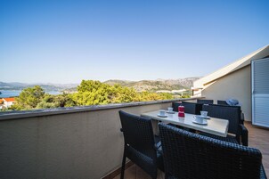 Un balcón con muebles de mimbre y una mesa, con vista a un paisaje montañoso y una masa de agua.