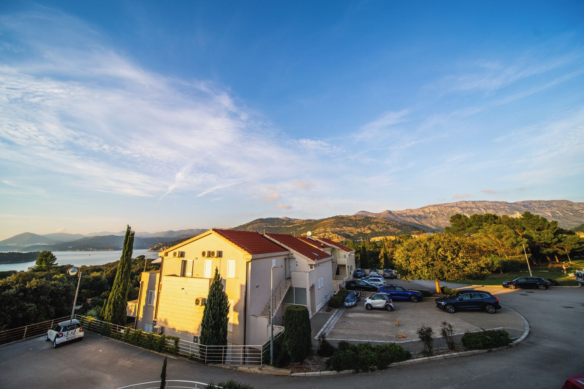 Um edifício com telhado vermelho, um estacionamento com vários carros, e uma vista de montanhas e um corpo d'água ao fundo.