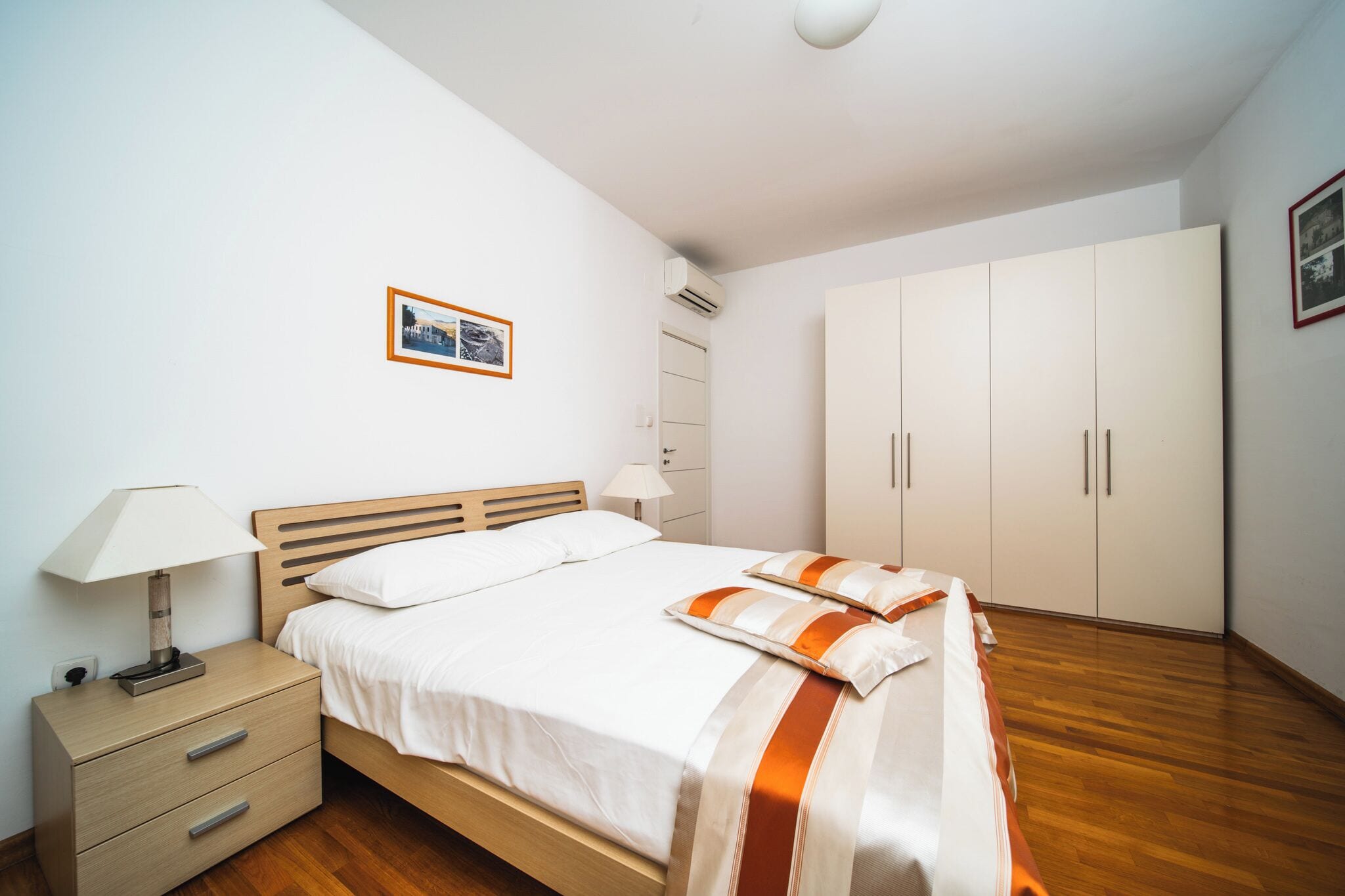 A bedroom with a wooden bed, white bedding, a nightstand with a lamp, a wardrobe, and a framed picture on the wall.
