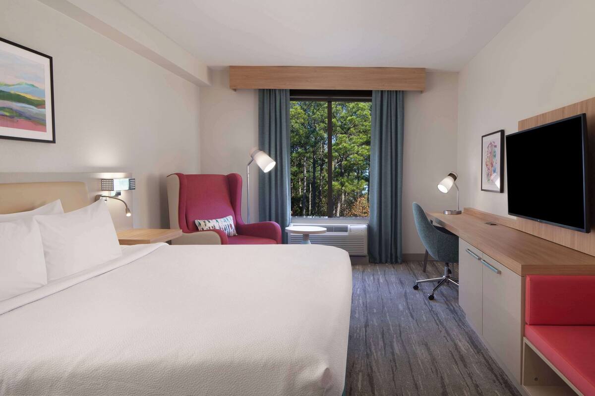 Une chambre d’hôtel avec un grand lit, un bureau avec une télévision, un fauteuil rouge et une fenêtre donnant sur des arbres.