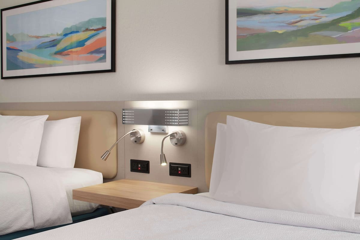 Une chambre d’hôtel avec deux lits, une table de chevet, une lampe fixée au mur et deux tableaux encadrés.