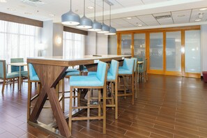 A modern hotel lobby with a wooden bar area, blue cushioned chairs, and a large window providing natural light. - Hampton Inn Garden City (Garden City)