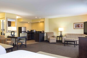 A hotel room with a bed, a desk, a chair, a sofa, a small kitchen area, and a television.