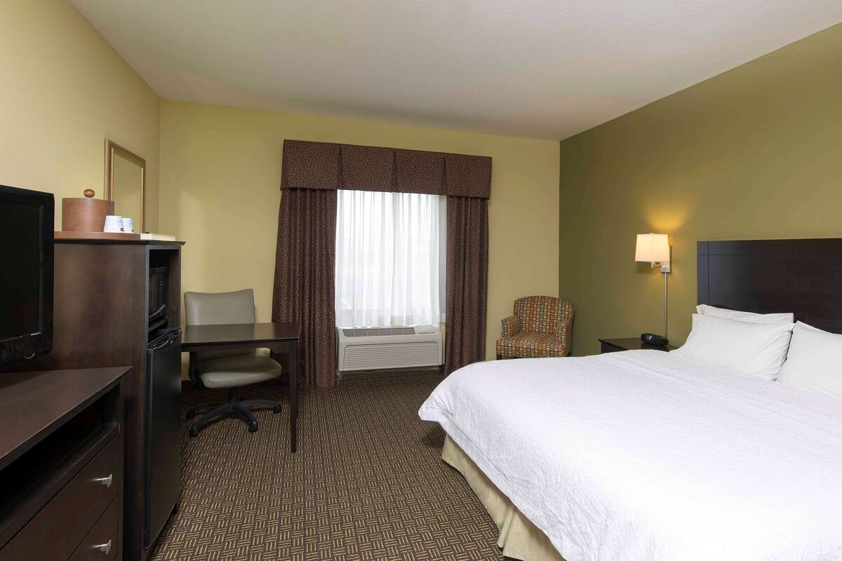 Una habitación de hotel con cama, escritorio, silla, televisor y ventana con cortinas.