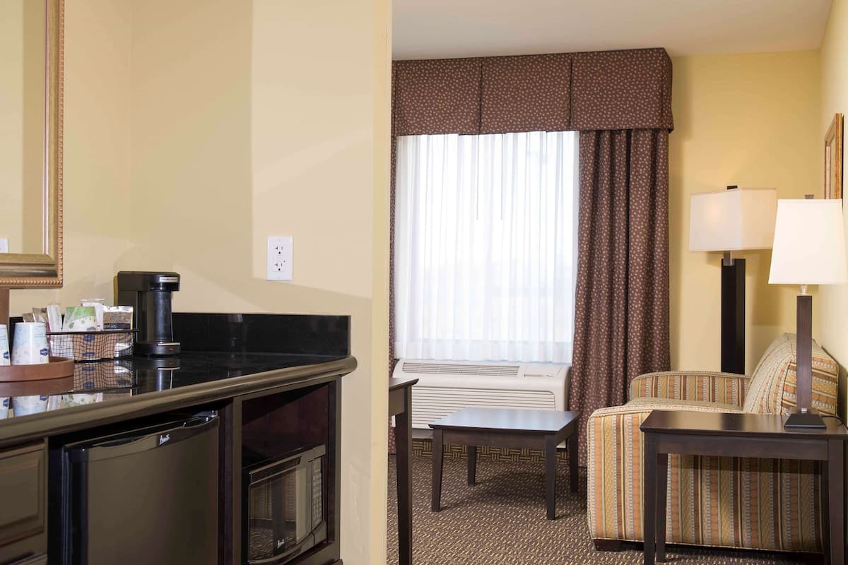 Una habitación de hotel con kitchenette, una mesa de comedor, un sofá y un amplio ventanal con cortinas.