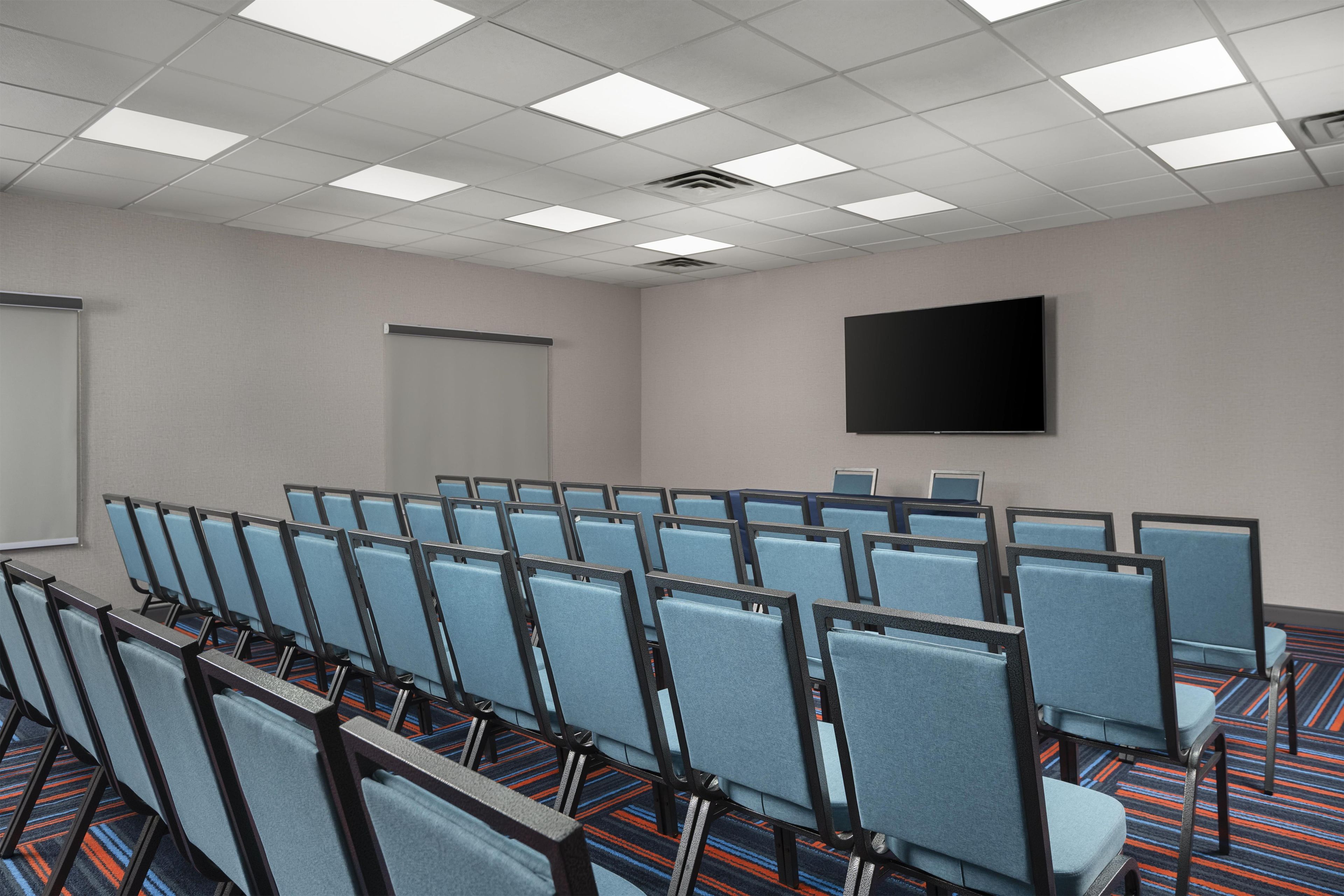 Une salle de conférence avec des rangées de chaises, un écran de projection et un téléviseur à écran plat fixé au mur.