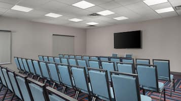 Une salle de conférence avec des rangées de chaises, un écran de projection et un téléviseur à écran plat fixé au mur.
