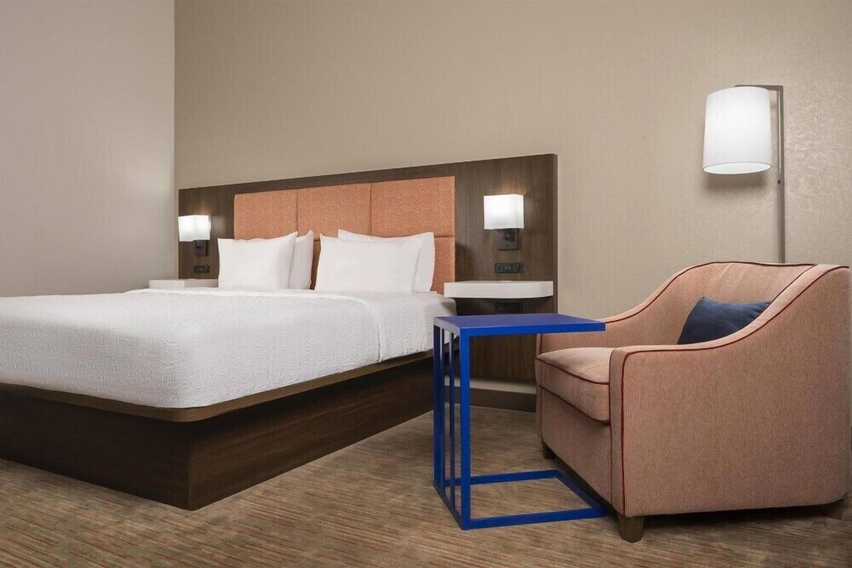 A hotel room with a bed, a chair, a small table, and two wall-mounted lamps.
