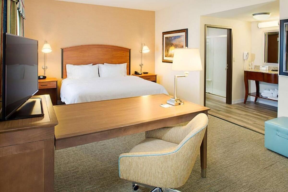 A hotel room with a bed, bedside lamps, a desk with a chair, a television, a desk lamp, a mirror, and a bathroom door.