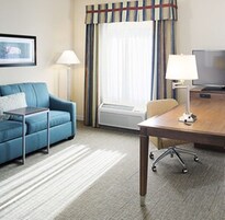 A hotel room with a blue sofa, a small table, a lamp, a TV, and a window with curtains.