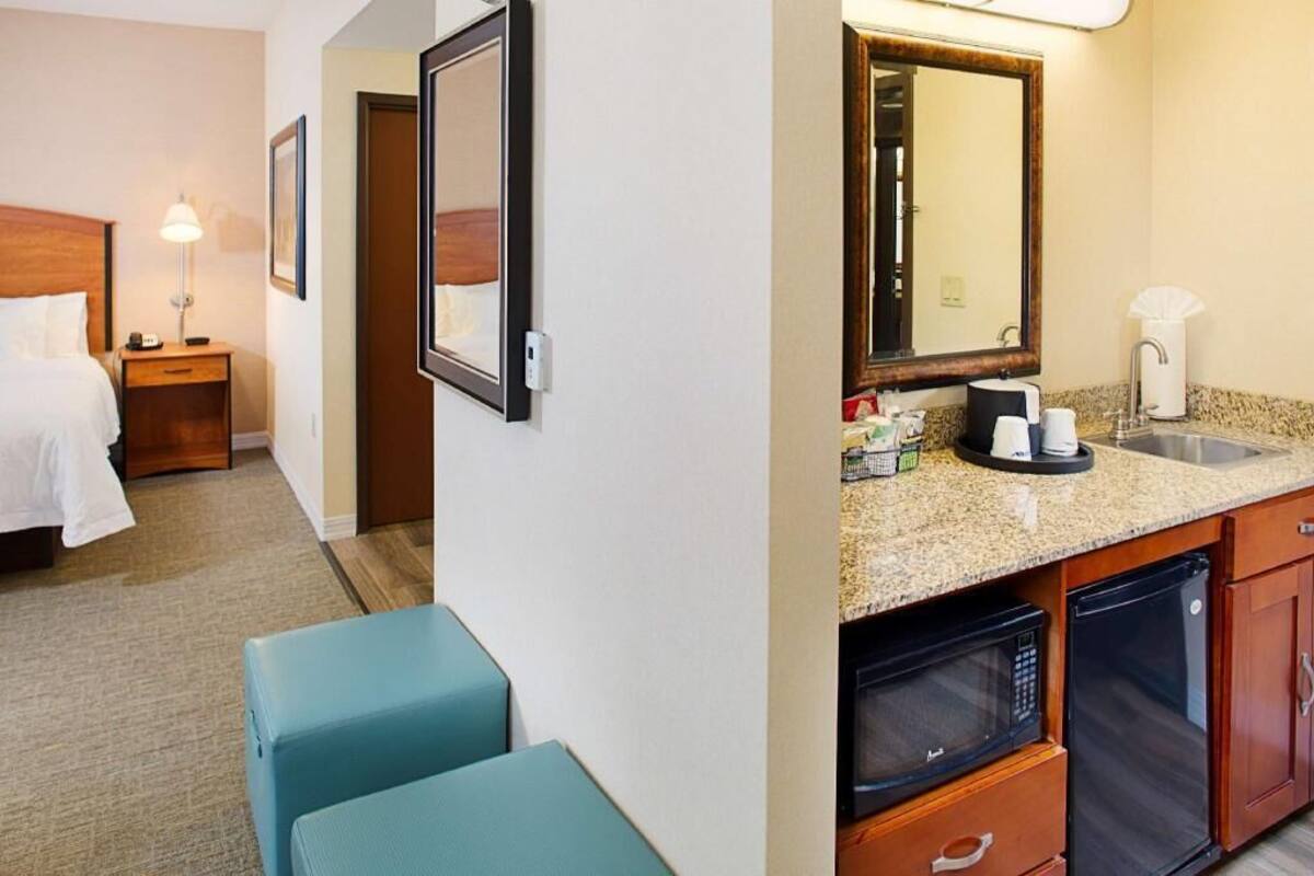 A hotel room with a bed, bedside table, lamp, mirror, kitchenette with microwave, refrigerator, and a seating area.