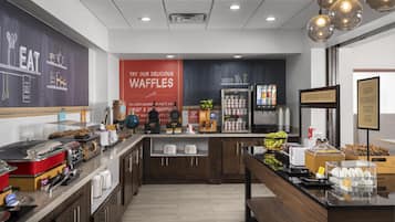 Un área de desayuno con una estación de waffles, máquinas de café y un refrigerador lleno de bebidas.