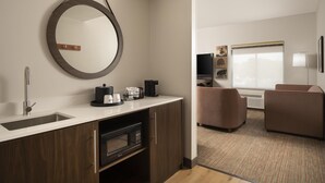 A modern hotel room with a kitchen area, a large mirror, a flat-screen TV, and a seating area with a sofa and armchair.