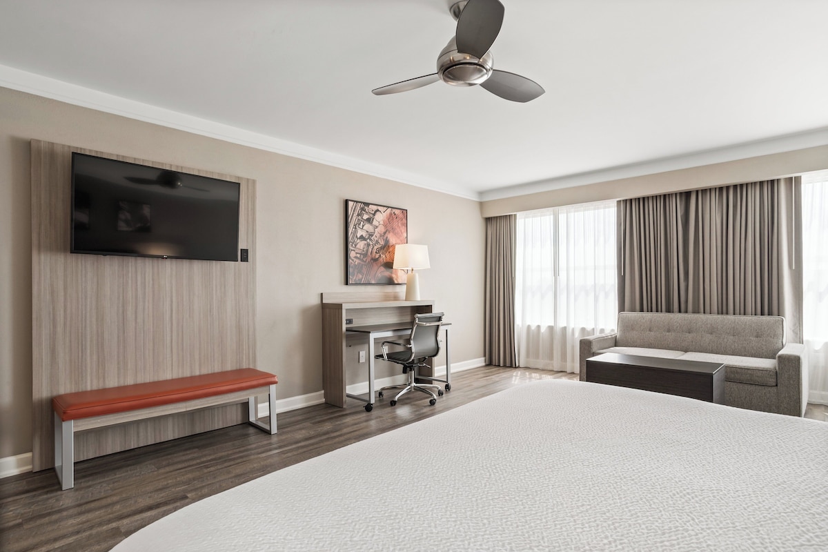 Ein Hotelzimmer mit einem großen Bett, einem Schreibtisch mit Stuhl, einer Bank, einem an der Wand befestigten Fernseher, einem Deckenventilator und einem Fenster mit Vorhängen.