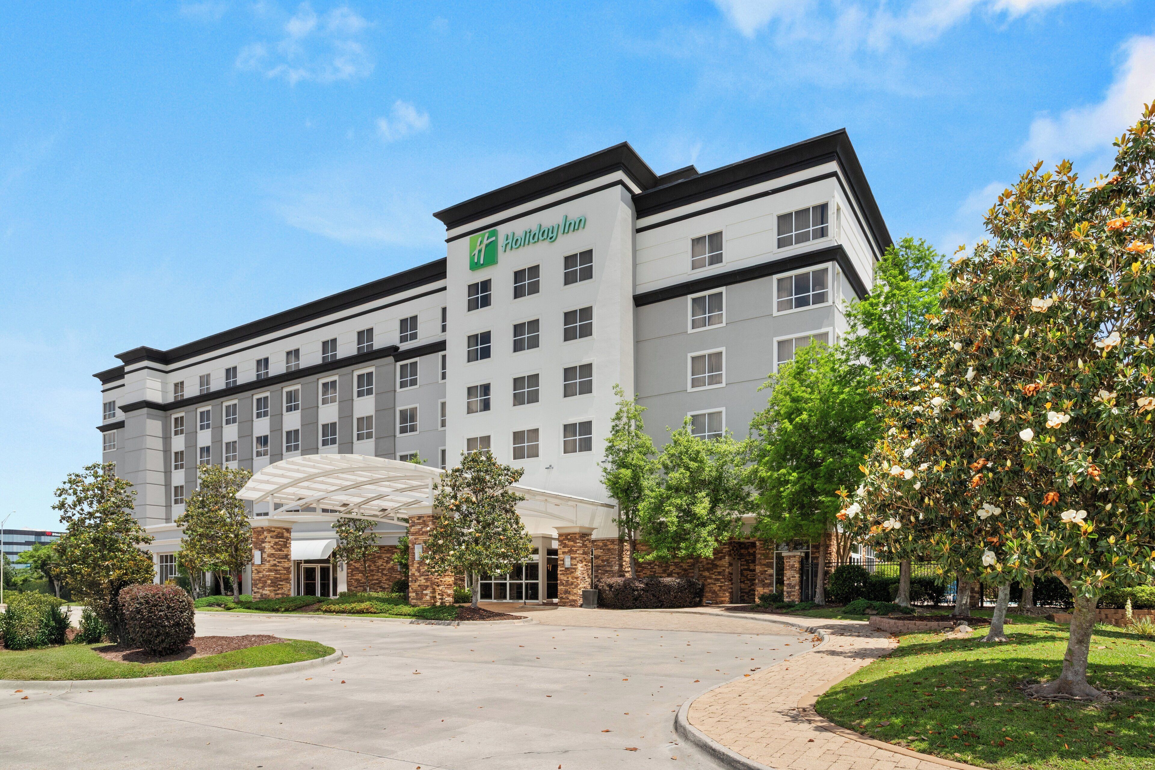 A Holiday Inn hotel with a modern design, featuring a central entrance with a glass canopy, surrounded by landscaped gardens and mature trees.