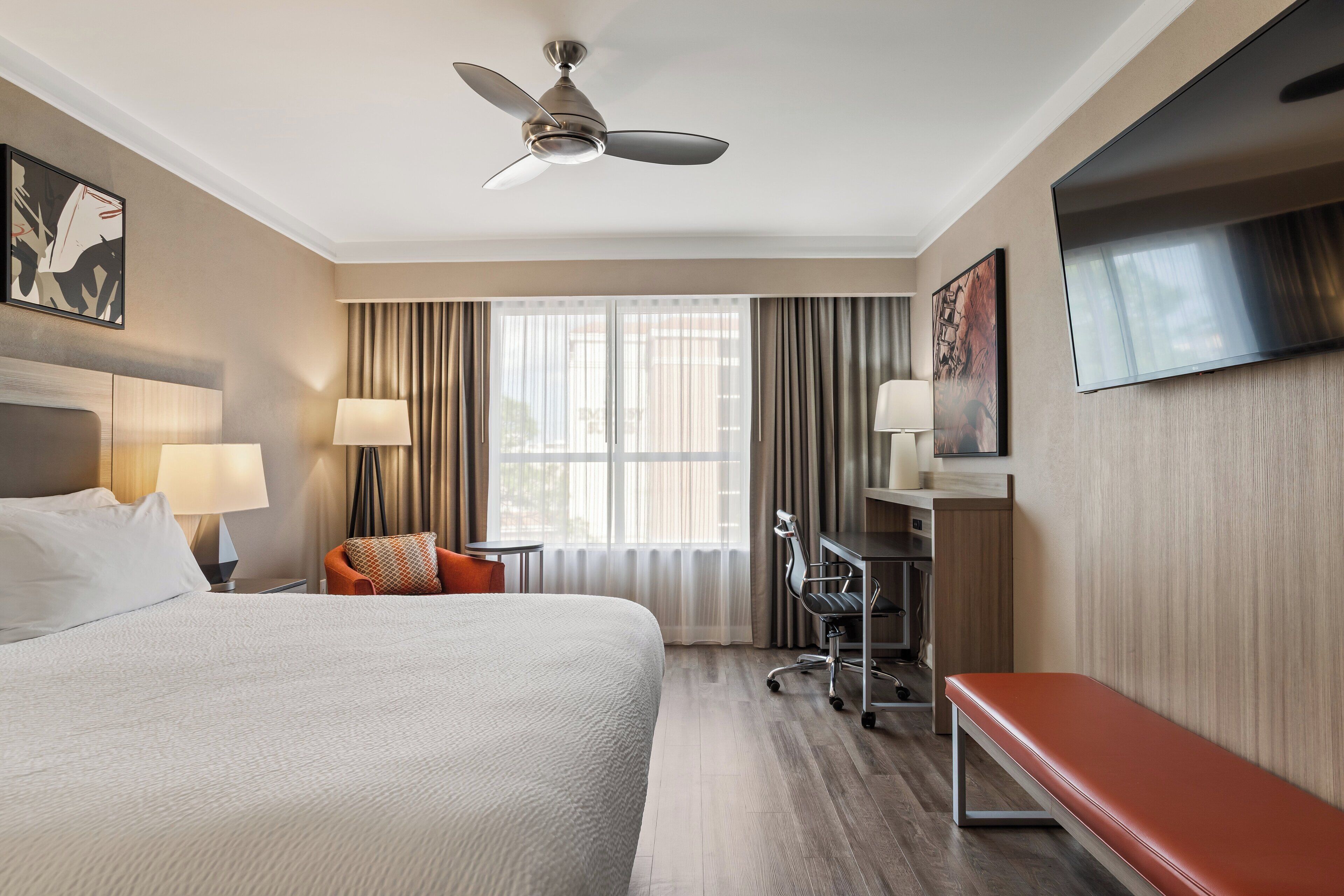 Een hotelkamer met een groot bed, een bureau met stoel, een aan de muur bevestigde televisie en een plafondventilator.