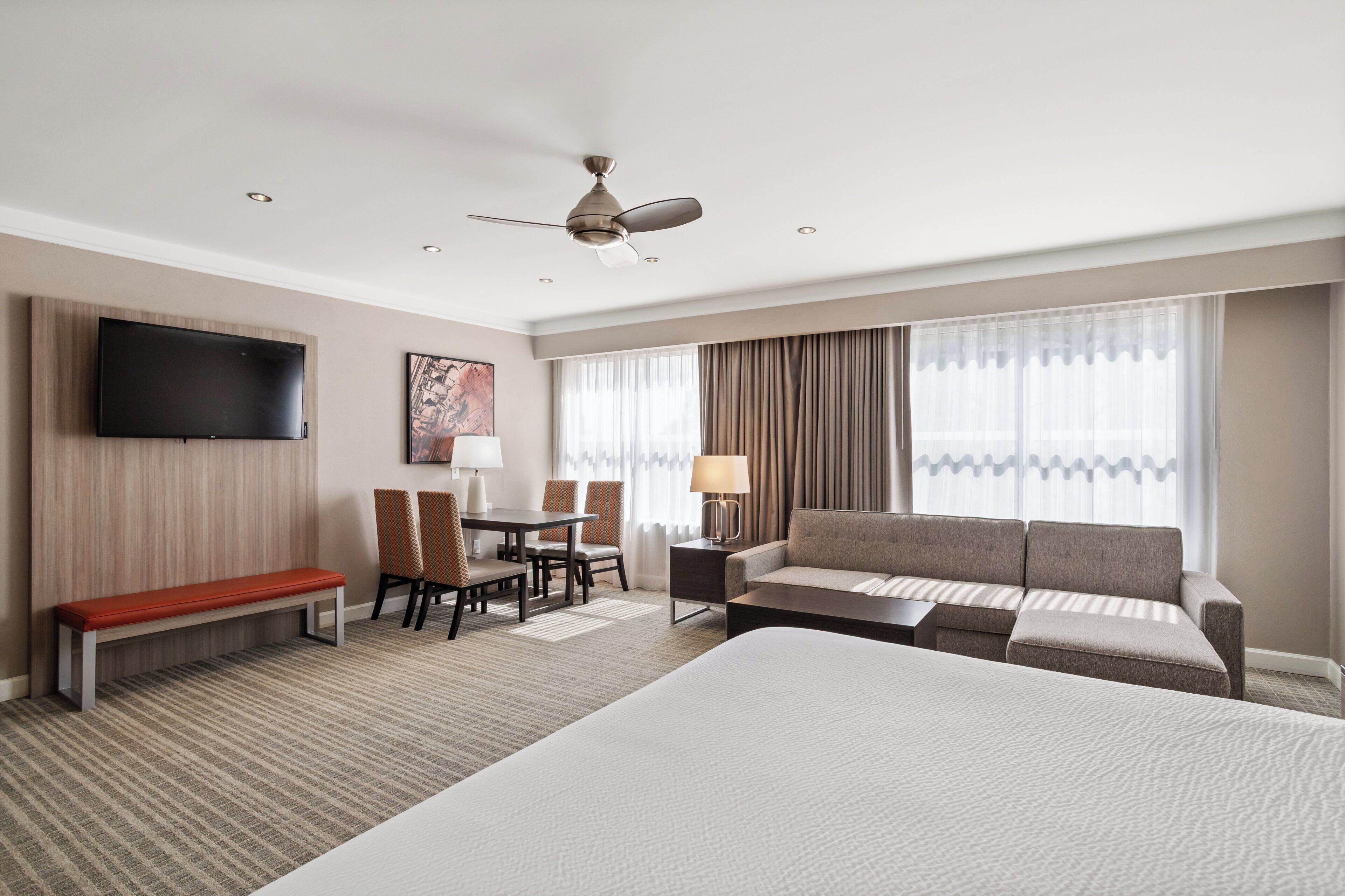 A hotel room with a large bed, a sofa, a desk with chairs, a TV mounted on the wall, and a ceiling fan.
