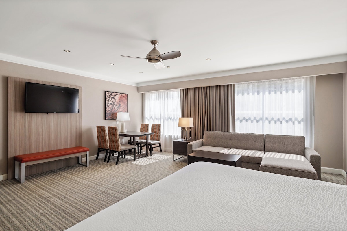 Ein Hotelzimmer mit einem großen Bett, einem Sofa, einem Schreibtisch mit Stühlen, einem an der Wand befestigten Fernseher und einem Deckenventilator.