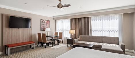 A hotel room with a large bed, a sofa, a desk with chairs, a TV mounted on the wall, and a ceiling fan.