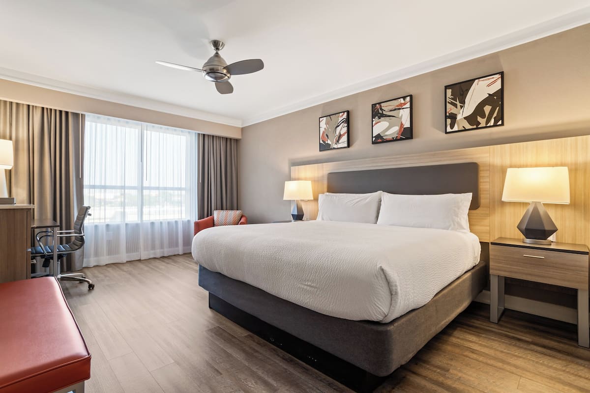 A hotel room with a large bed, two bedside tables with lamps, a ceiling fan, a desk with a chair, and a window with curtains.