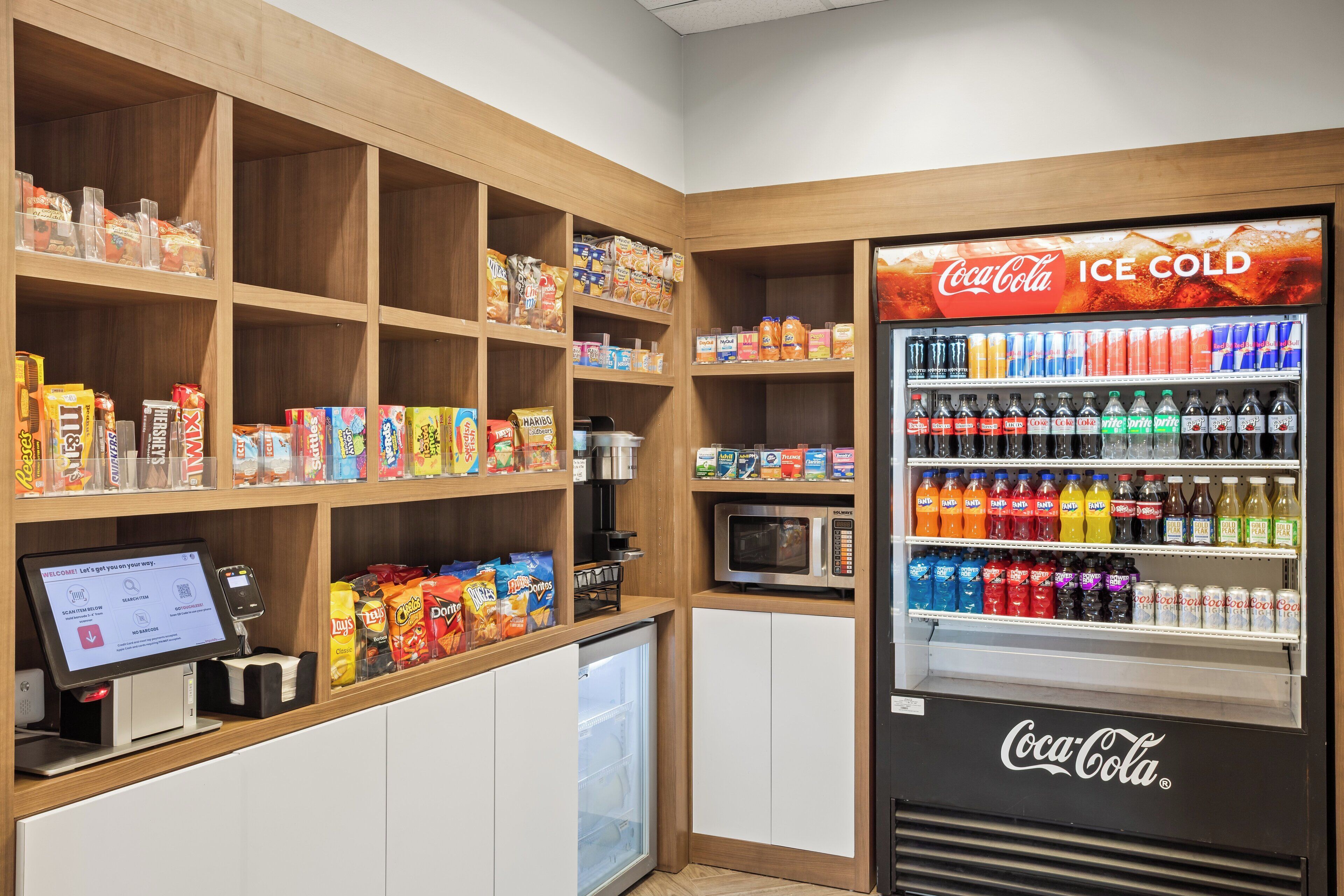 A self-service kiosk with a Coca-Cola refrigerator stocked with various beverages, a microwave, and shelves with snacks and food items.