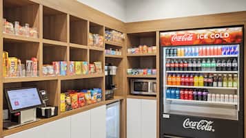 A self-service kiosk with a Coca-Cola refrigerator stocked with various beverages, a microwave, and shelves with snacks and food items.