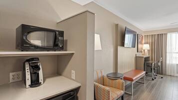 A hotel room with a kitchenette, a microwave, a coffee maker, a TV, a chair, a table, and a lamp.